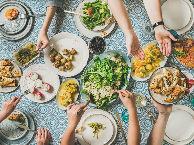 Mediterranean style dinner. Flat-lay of table with salads, starters, pastries over blue table cloth with hands holding drinks, sharing food, top view. Holiday gathering, vegetarian party concept