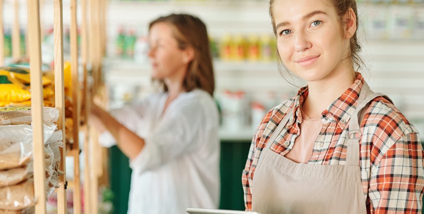 Young pretty female assistant of gardening supermarket in workwear using digital tablet while searching for goods in their online shop